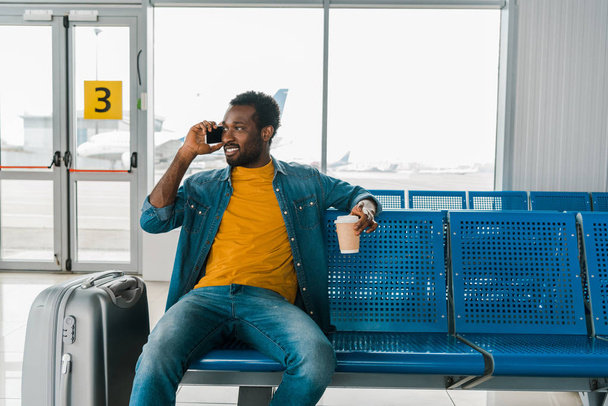smiling african american man sitting in departure lounge with coffee to go and talking on smartphone - Фото, изображение