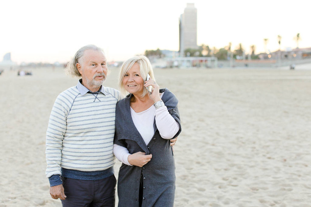 Starší manžel chodit na písčité pláži s blond ženy mluví o smartphone. - Fotografie, Obrázek