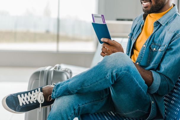 bijgesneden weergave van glimlachende Afro-Amerikaanse man zittend in de luchthaven met vliegticket en paspoort - Foto, afbeelding