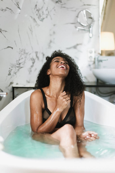 風呂に座っている若いかわいいアフロアメリカンガール, 黒い水着を着て. - 写真・画像