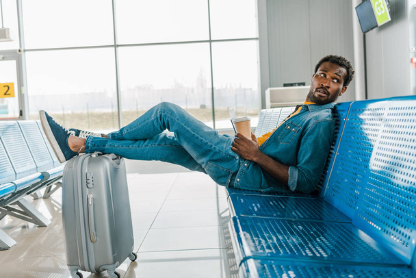 homem americano africano sentado no aeroporto com café para ir e pernas na mala
 - Foto, Imagem