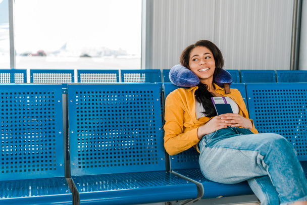 усміхнена афроамериканська жінка сидить з подушкою для подорожей та паспортом та авіаквитком у залі вильоту
 - Фото, зображення