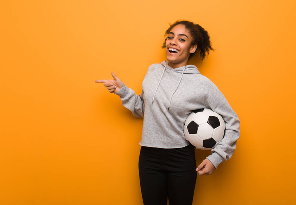 Молодая фитнес черная женщина указывает пальцем в сторону. Холдинг футбольный мяч
. - Фото, изображение