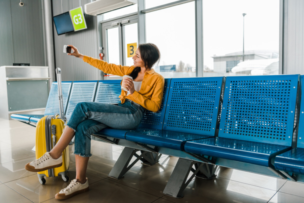 Smiling Afro-Amerikaanse vrouw zittend in vertrek lounge met koffer, koffie te gaan en rekening selfie op smartphone - Foto, afbeelding