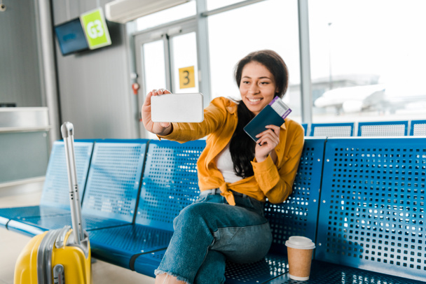 Happy Afro-Amerikaanse vrouw zit in vertrek lounge met koffer, koffie te gaan en rekening selfie met paspoort en vliegticket op smartphone - Foto, afbeelding