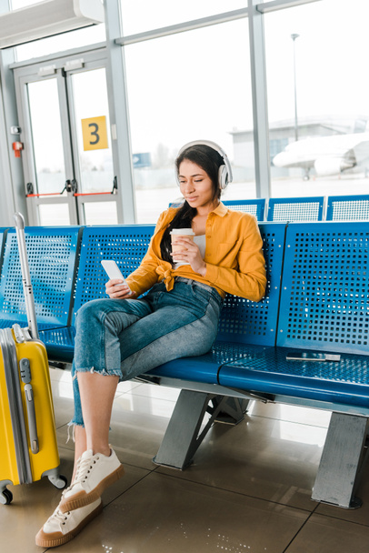 feliz mulher afro-americana sentada na sala de partida com café para ir e mala enquanto ouve música em fones de ouvido e usando smartphone
 - Foto, Imagem