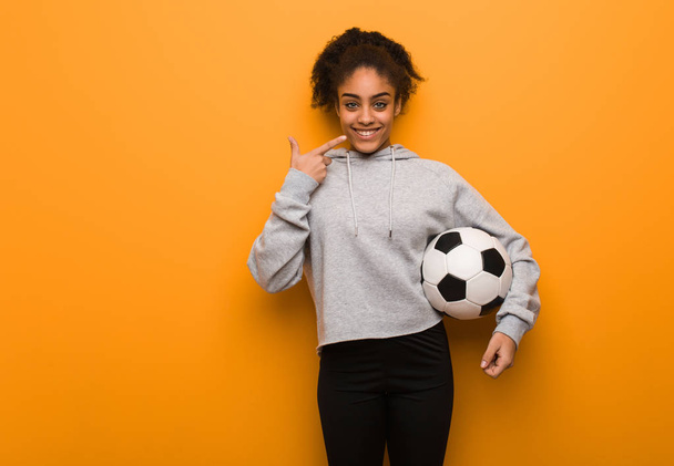 Młoda fitness czarna kobieta uśmiecha się, wskazując usta. Trzymanie piłki nożnej. - Zdjęcie, obraz