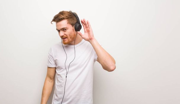 Молодой рыжий человек пытается слушать сплетни. Слушать музыку в наушниках
. - Фото, изображение