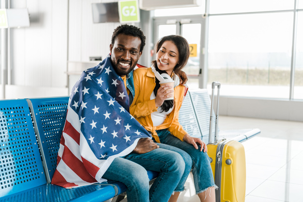 feliz pareja afroamericana sentada con bandera americana y equipaje en la sala de salida en el aeropuerto
  - Foto, imagen