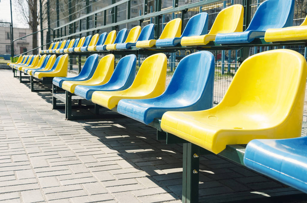 leere Reihen blauer und gelber Sitze auf dem Schulsportplatz, im Freien - Foto, Bild