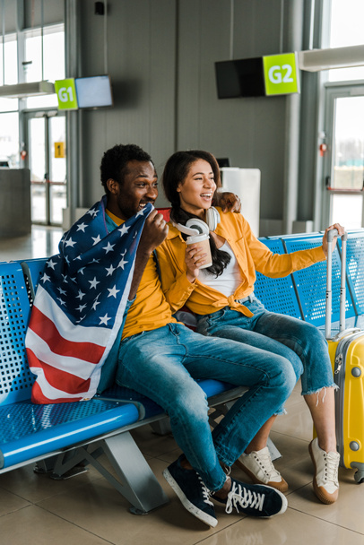 空港の出発ラウンジでアメリカ国旗と荷物で座って幸せなアフリカ系アメリカ人のカップル  - 写真・画像