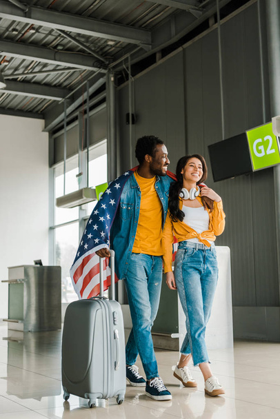 空港の出発ラウンジで歩くアメリカ国旗とスーツケースとの幸せなアフリカ系アメリカ人のカップル  - 写真・画像