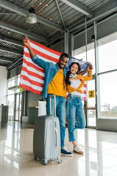 空港の出発ラウンジで歩くアメリカの国旗と荷物を持つアフリカ系アメリカ人のカップルの笑顔  - 写真・画像