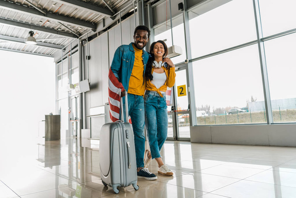 Happy Afro-Amerikaanse paar met Amerikaanse vlag en bagage wandelen in vertrek lounge in de luchthaven  - Foto, afbeelding