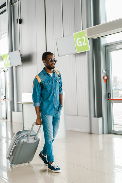 サングラスでハンサムなアフリカ系アメリカ人の男性を幸せにする空港でスーツケースを歩いて - 写真・画像