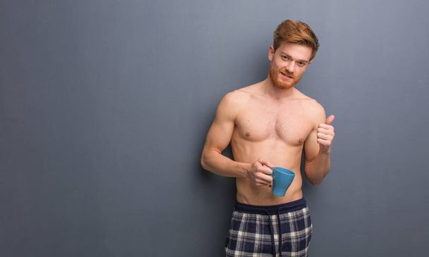 Young shirtless redhead man smiling and raising thumb up. He is holding a coffee mug. - Photo, Image