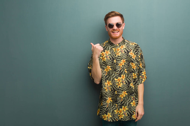 junger rothaariger Mann in exotischer Sommerkleidung, der mit dem Finger zur Seite zeigt - Foto, Bild