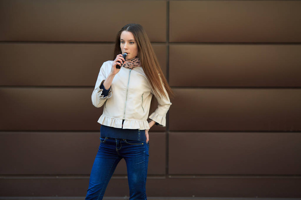 Vape teenager. Young pretty white girl in casual clothing smoking an electronic cigarette opposite modern brown background on the street in the spring. Bad habit. Vaping activity. - Fotografie, Obrázek
