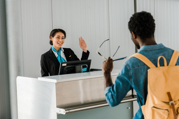 selektywne skupienie uśmiechnięty African American Airport robotnik machając ręką do turysty z plecaka - Zdjęcie, obraz