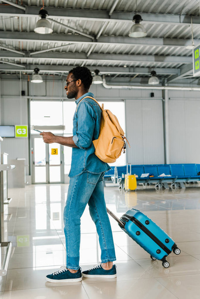 空港でスーツケースとバックパックとデニムの衣装で陽気なアフリカ系アメリカ人の男 - 写真・画像