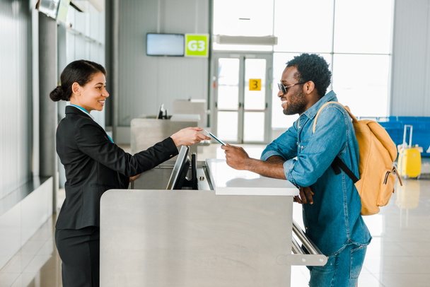 souriant travailleur afro-américain de l'aéroport prenant passeport et billet d'avion du touriste avec sac à dos
 - Photo, image