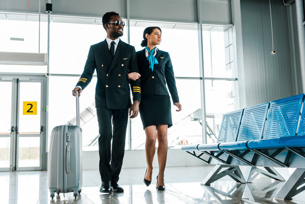 african american pilot and stewardess walking together with suitcase in airport - 写真・画像