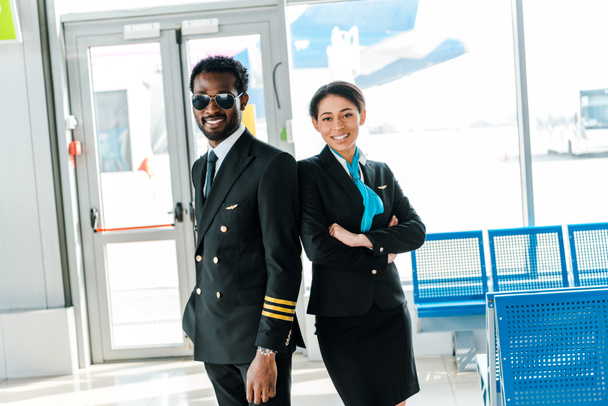空港で一緒に立って腕を組んでサングラスとスチュワーデスに微笑むアフリカ系アメリカ人のパイロット - 写真・画像
