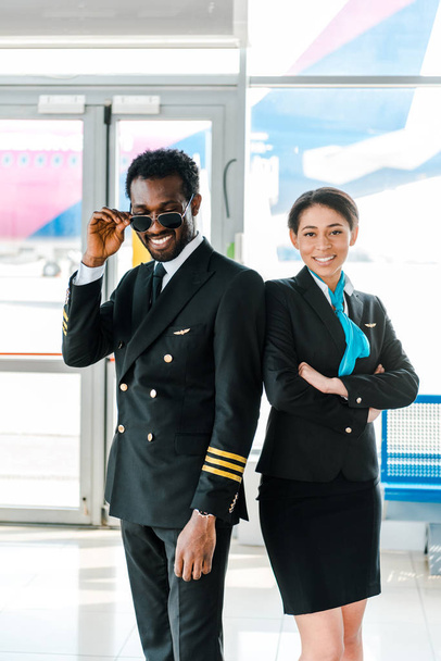 pilote afro-américain en lunettes de soleil et hôtesse de l'air avec les bras croisés posant ensemble à l'aéroport
 - Photo, image