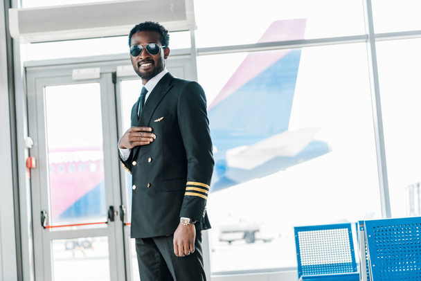 smiling african american pilot standing in departure lounge in airport - Photo, image