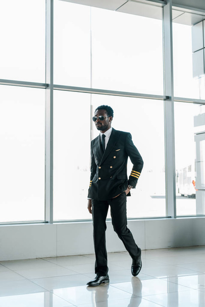 pilote afro-américain confiant dans des lunettes de soleil marchant à l'aéroport avec la main dans la poche
 - Photo, image