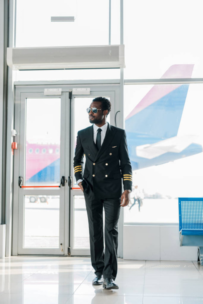 confident african american pilot in sunglasses walking in airport - Фото, изображение