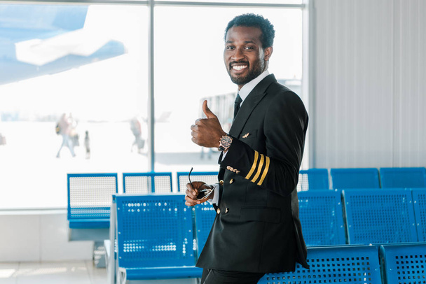smiling african american pilot showing thumb up in departure lounge  - Foto, afbeelding