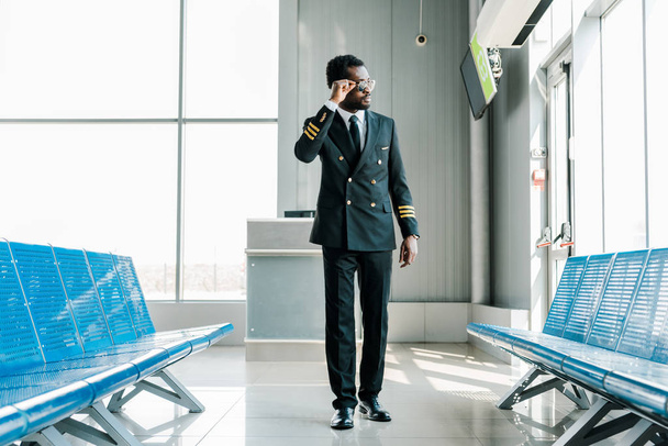 confident african american pilot in sunglasses walking in departure lounge in airport and looking at window - 写真・画像