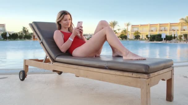 Sexy girl relaxing near the pool with a mobile phone. Woman texting and browsing the internet on mobile phone, lying on deck chair in hotel pool side area. Sunbathing and relaxing at resort. - Metraje, vídeo