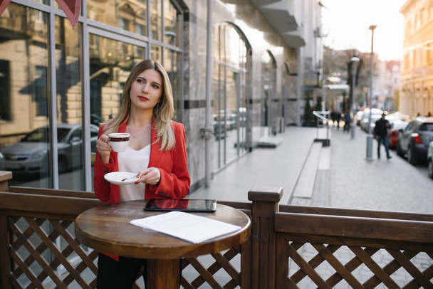 Beautiful young businesswoman with a disposable coffee cup, drinking coffee, and holding tablet in her hands against urban city background. - Фото, изображение