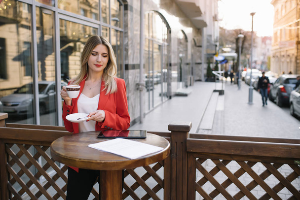 Beautiful young businesswoman with a disposable coffee cup, drinking coffee, and holding tablet in her hands against urban city background. - Fotó, kép