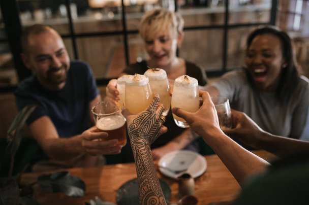 Laughing group of diverse friends toasting together with drinks while sitting together in a bar at night - Foto, immagini