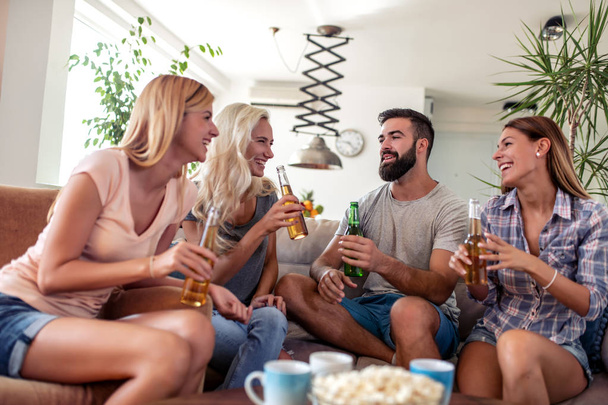 Ομάδα νεαρών φίλων που διασκεδάζουν, πίνοντας μπίρες και τρώγοντας ποπ κορν στο σπίτι. - Φωτογραφία, εικόνα