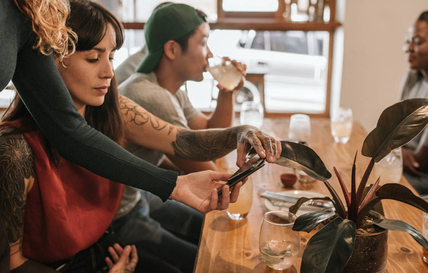 Young customer with tattoos using her cellphone and an nfc terminal to pay for the check in restaurant while out with friends - 写真・画像