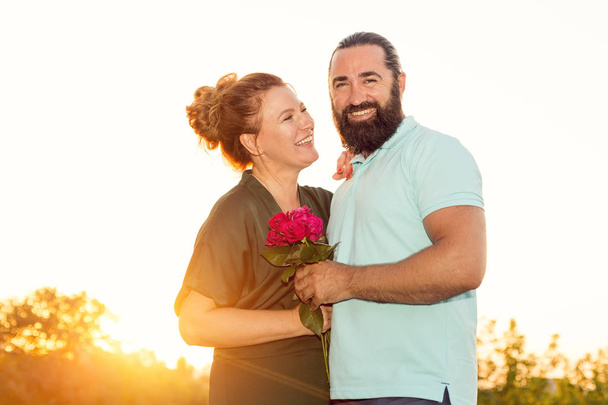 Happy middle-aged couple in love on sunset background. A woman with a bouquet of roses hugs her man. - Foto, Bild