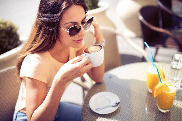 Young pretty woman drinking cappuccino, coffee in cafe outdoors, enjoying sunny morning in the city. - Fotografie, Obrázek