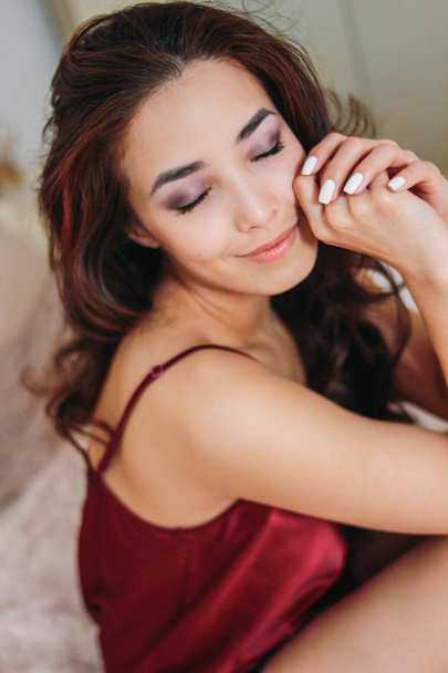 glücklich sinnlich lächelnde Mädchen asiatische junge Frau mit dunklen langen lockigen Haaren in roter Unterwäsche auf dem Bett sitzend - Foto, Bild