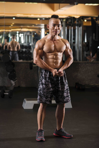 Χαμογελαστός όμορφος Βιετναμέζος bodybuilder κάνει πόζα στήθους μπροστά από τον καθρέφτη - Φωτογραφία, εικόνα