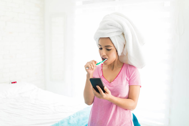 Frau mit Handtuch umwickelt auf dem Kopf Zähne putzen und mit Anwendungen auf dem Handy im Schlafzimmer - Foto, Bild