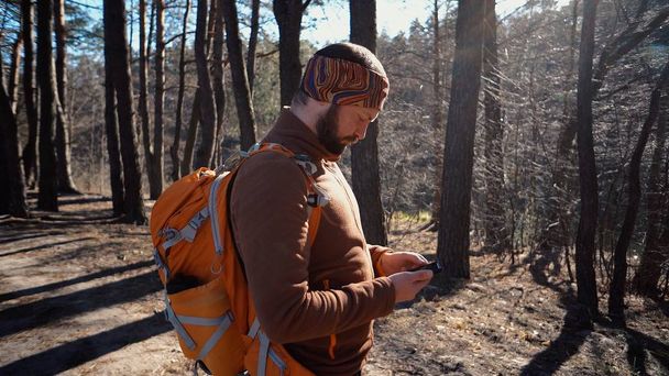 theme tourism and technology. Young caucasian man with beard and backpack. Hiking tourist in pine forest uses technology, hand holding mobile phone to touch the screen. Gps application orientation - Photo, Image