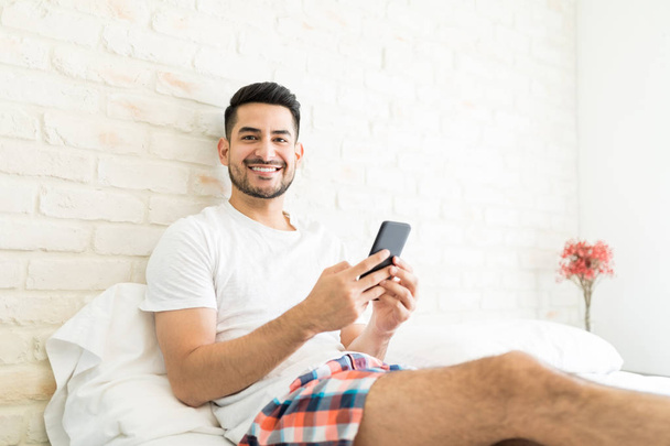 Портрет привлекательного мужчины, устанавливающего новое приложение на смартфон в спальне
 - Фото, изображение