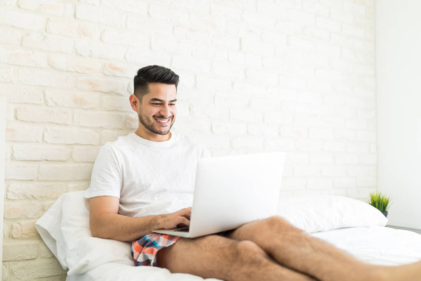 Щасливий молодий чоловік відпочиває на зручному ліжку і спілкується в соціальних мережах з найкращим другом на ноутбуці
 - Фото, зображення