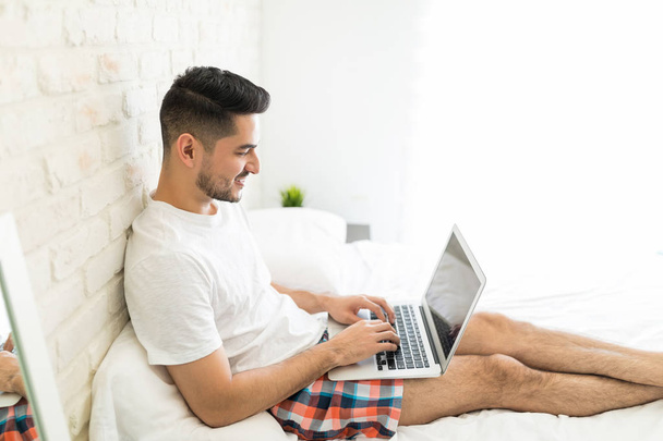 Mosolygós fiatalember használó laptop alatt szabadidejükben otthon - Fotó, kép