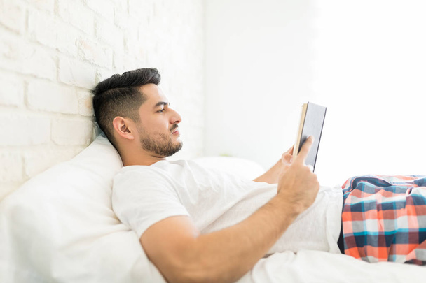 Vista lateral del hombre guapo leyendo libro mientras está acostado en la cama
 - Foto, Imagen