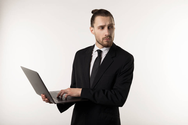 Vonzó szakállas középvezető visel fekete öltönyt és a nyakkendő gondolkodás laptop a kezében, hogyan lehet megoldani a problémát okozta a főnök izolált fehér háttér - Fotó, kép
